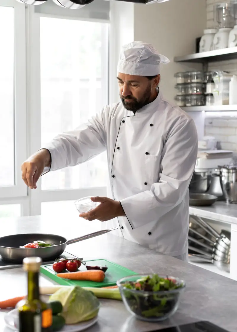 chef in white uniform seasoning food in a pan, with fresh vegetables on the counter