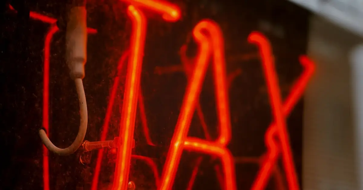 A-glowing-red-neon-sign-spelling-_TAX_-reflected-on-a-glass-surface-set-against-a-blurred-background-of-trees-and-power-lines