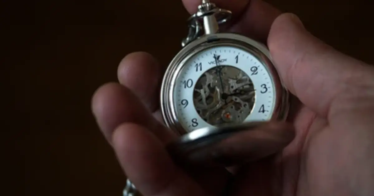 A-person-holds-a-silver-pocket-watch-revealing-intricate-gears-and-a-clear-face-with-black-numbers-against-a-dark-background