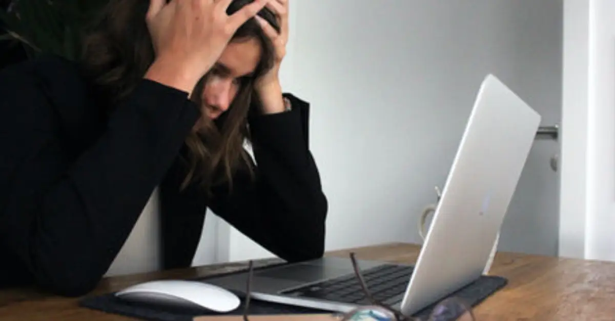 A-person-in-a-black-blazer-sits-at-a-desk-hunched-over-a-laptop-appearing-stressed-with-hands-on-their-head