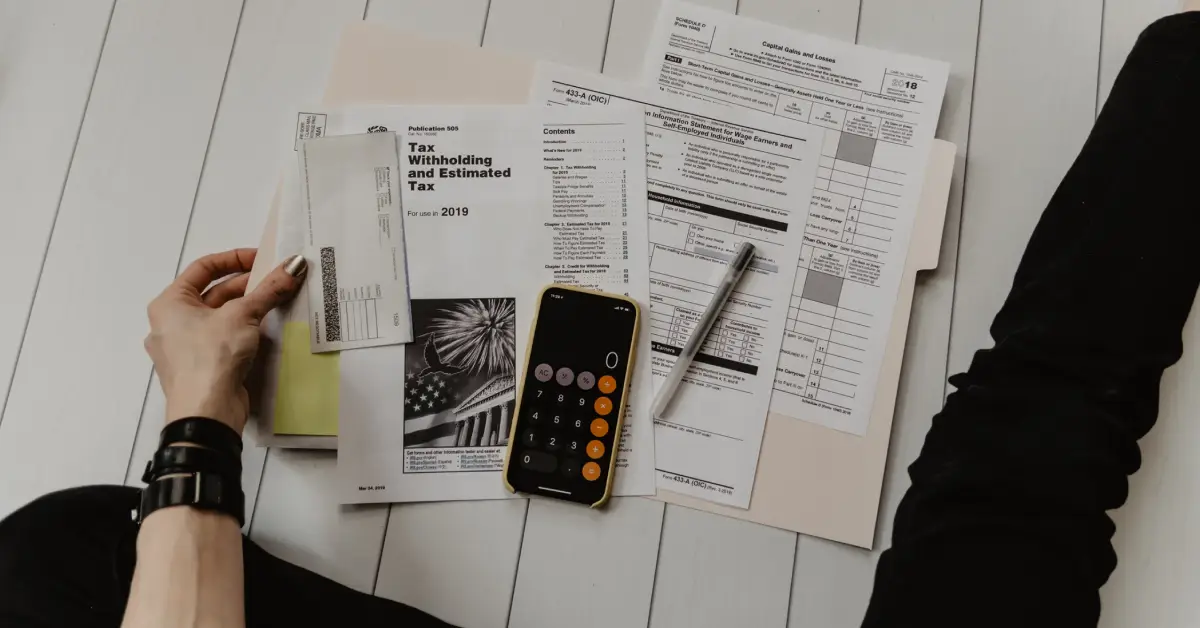 An-individual-organizing-tax-documents-and-forms-on-a-light-wooden-surface-featuring-a-calculator-and-a-handwritten-note