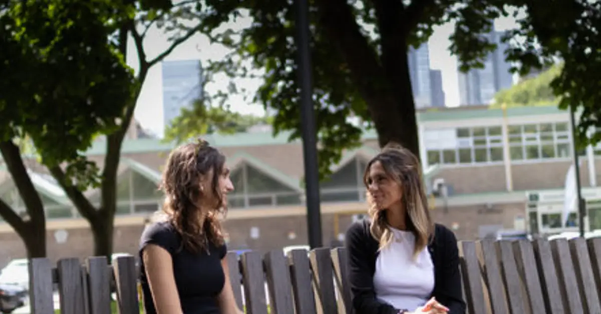 Two-women-sit-on-a-wooden-bench-in-a-park-engaging-in-conversation-surrounded-by-trees-and-a-cityscape-in-the-background